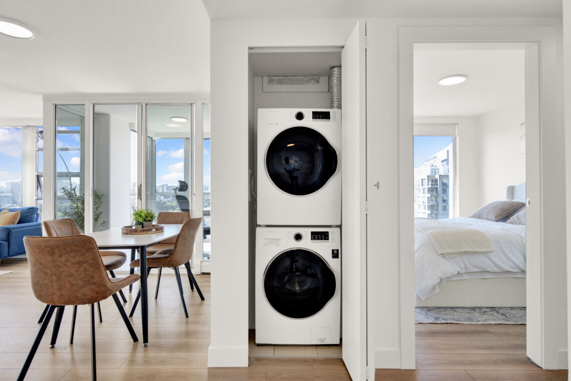 Open laundry closet with stacked front-loading washer and dryer, flanked by doors in a bright open-plan apartment with dining area to the left and a bedroom to the right.