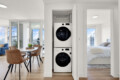 Open laundry closet with stacked front-loading washer and dryer, flanked by doors in a bright open-plan apartment with dining area to the left and a bedroom to the right.