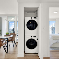 Open laundry closet with stacked front-loading washer and dryer, flanked by doors in a bright open-plan apartment with dining area to the left and a bedroom to the right.