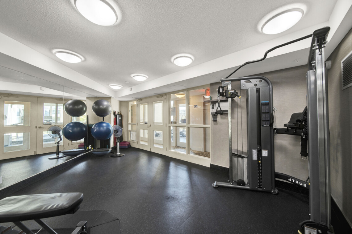 Gym workout room with a large mirrored wall, exercise machines on the right, and stability balls near the windows.