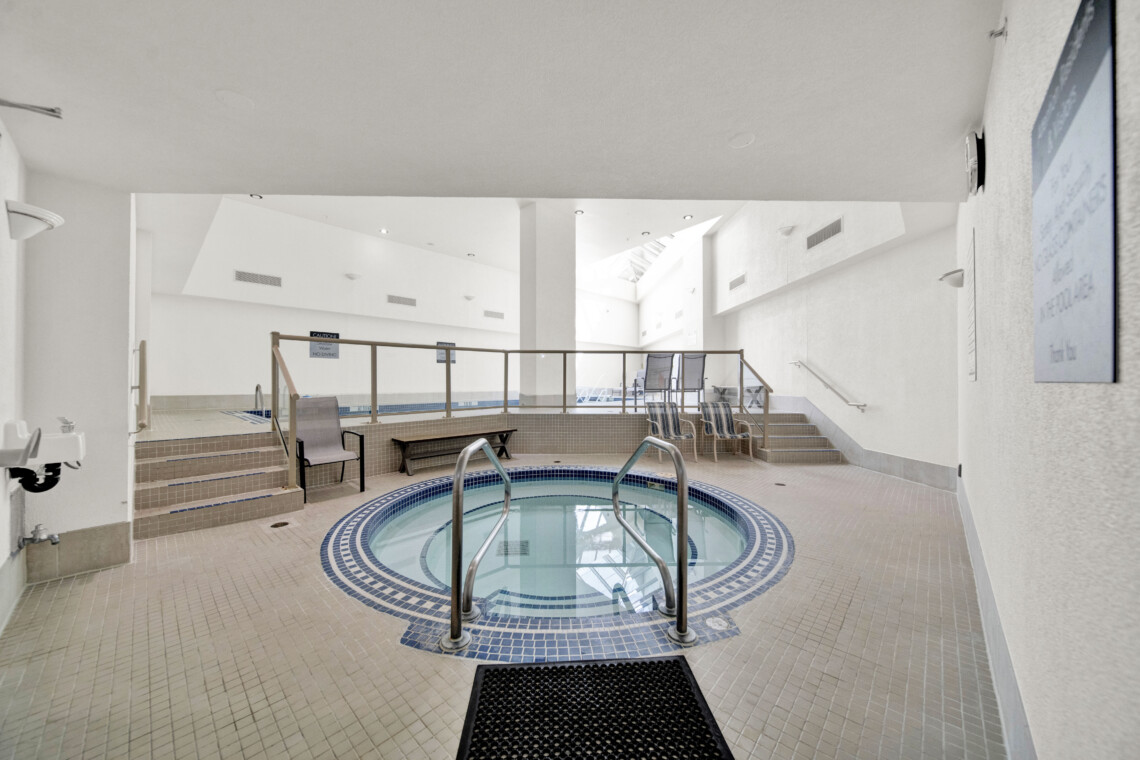 Indoor pool area with a circular hot tub in the foreground, tiled floor, and white walls; seating and railings in the background.