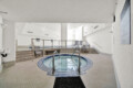 Indoor pool area with a circular hot tub in the foreground, tiled floor, and white walls; seating and railings in the background.