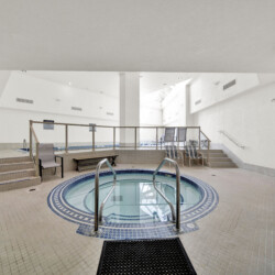 Indoor pool area with a circular hot tub in the foreground, tiled floor, and white walls; seating and railings in the background.