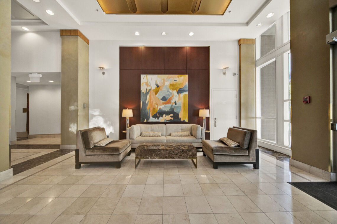 Lobby seating area with beige sofas, a colorful abstract painting centered on a dark wood wall, and two table lamps flanking the artwork in a bright lobby.