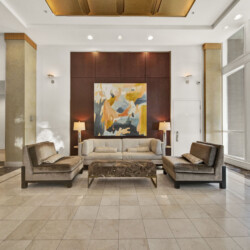 Lobby seating area with beige sofas, a colorful abstract painting centered on a dark wood wall, and two table lamps flanking the artwork in a bright lobby.