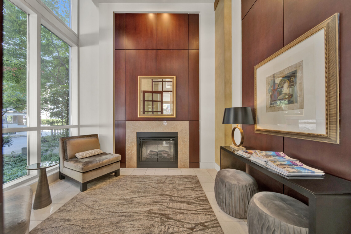 Elegant living room lobby with a wood-paneled wall, central fireplace, framed mirror, and seating arrangement with ottomans and chairs, low light glowing above.