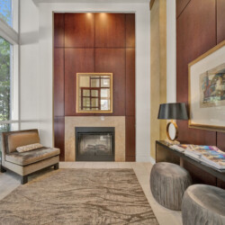 Elegant living room lobby with a wood-paneled wall, central fireplace, framed mirror, and seating arrangement with ottomans and chairs, low light glowing above.