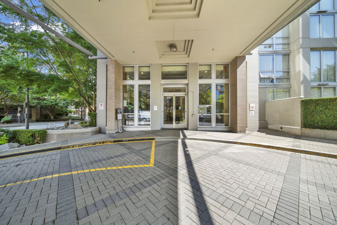 Entrance to a modern building with glass doors under a wide concrete canopy, brick columns, and yellow pickup/drop-off curb lines in the foreground.