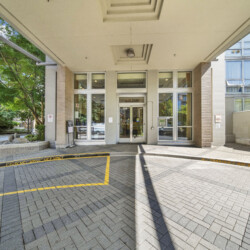 Entrance to a modern building with glass doors under a wide concrete canopy, brick columns, and yellow pickup/drop-off curb lines in the foreground.