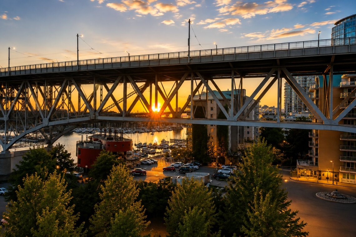 Sunset scene with a steel truss bridge over a marina, boats in the water, and city buildings in the background. Trees frame the foreground.