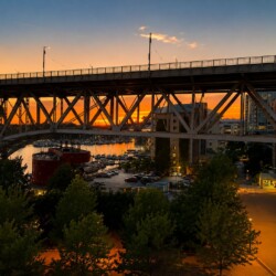 Sunset over a large metal bridge spanning a marina with yachts and orange sky in the background, city buildings on the right.