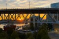Sunset behind a steel bridge over a marina, with boats, cars, and city buildings visible in the distance, beneath a golden sky.