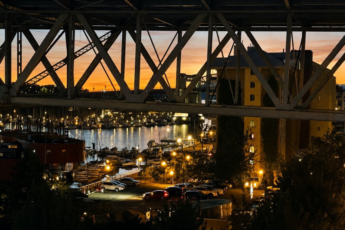 Harbor at sunset viewed under a large steel bridge, with moored boats and a lit waterfront.