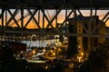 Harbor at sunset viewed under a large steel bridge, with moored boats and a lit waterfront.