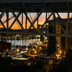 Harbor at sunset viewed under a large steel bridge, with moored boats and a lit waterfront.
