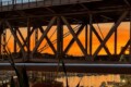 Sunset over a metal truss bridge spanning a harbor, with calm water reflecting lights and boats below.