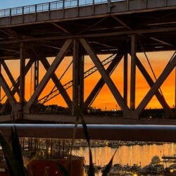 Sunset over a metal truss bridge spanning a harbor, with calm water reflecting lights and boats below.