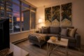 Cozy living room with a gray sectional, throw blanket, and pillows, lit by a floor lamp, facing a city view through large windows.