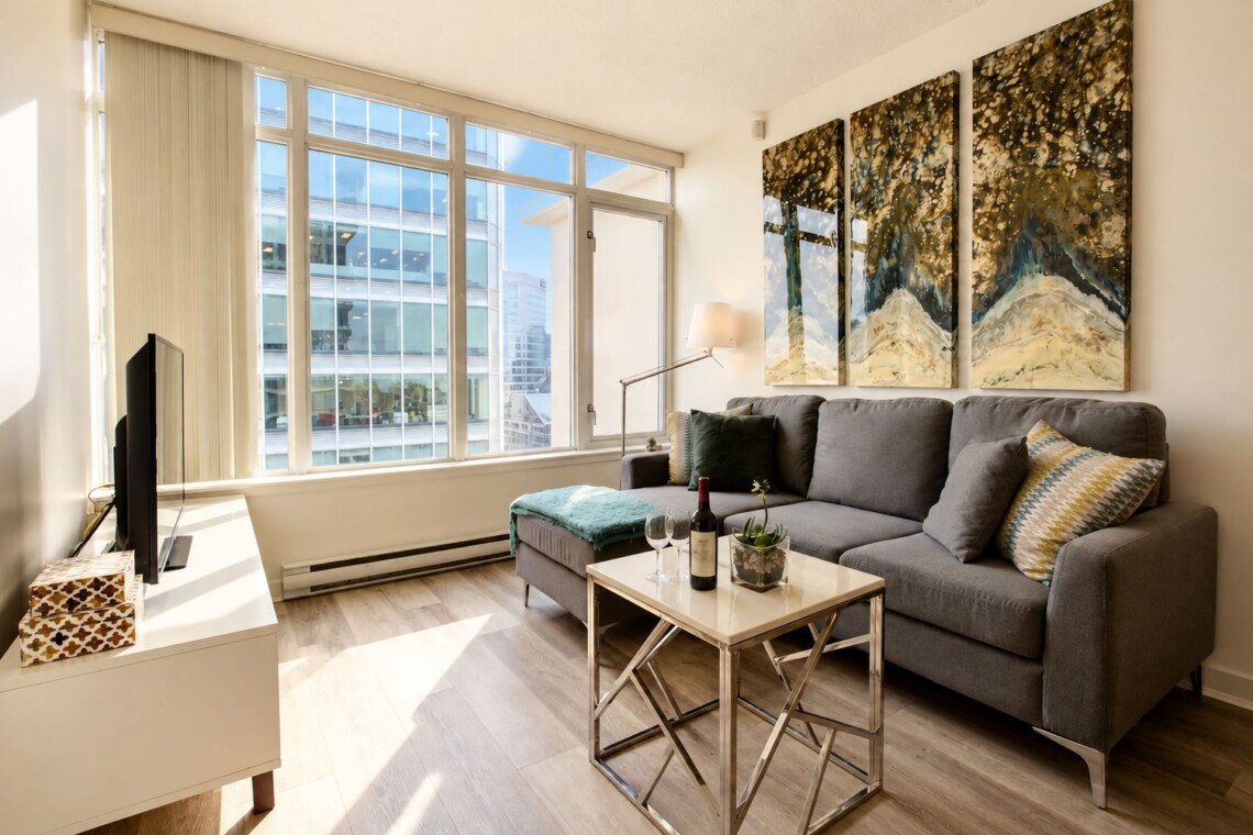 Bright living room with tall windows letting in sunlight, a grey sofa, and a metal-framed coffee table; three-panel abstract artwork hangs on the wall above the sofa.