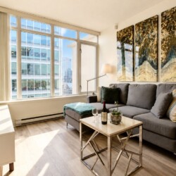 Bright living room with tall windows letting in sunlight, a grey sofa, and a metal-framed coffee table; three-panel abstract artwork hangs on the wall above the sofa.