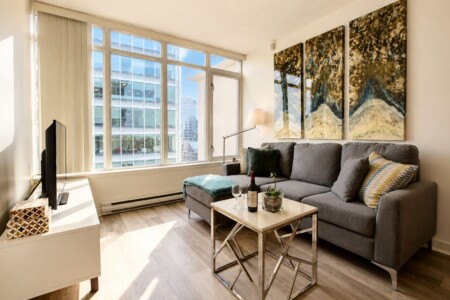 Bright living room with tall windows letting in sunlight, a grey sofa, and a metal-framed coffee table; three-panel abstract artwork hangs on the wall above the sofa.