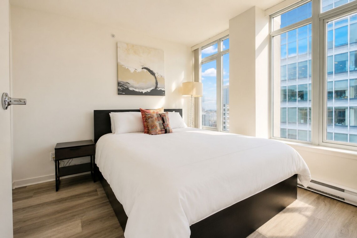 Bright hotel bedroom with a white bed, black wood headboard, and a small dark nightstand under a large abstract painting.