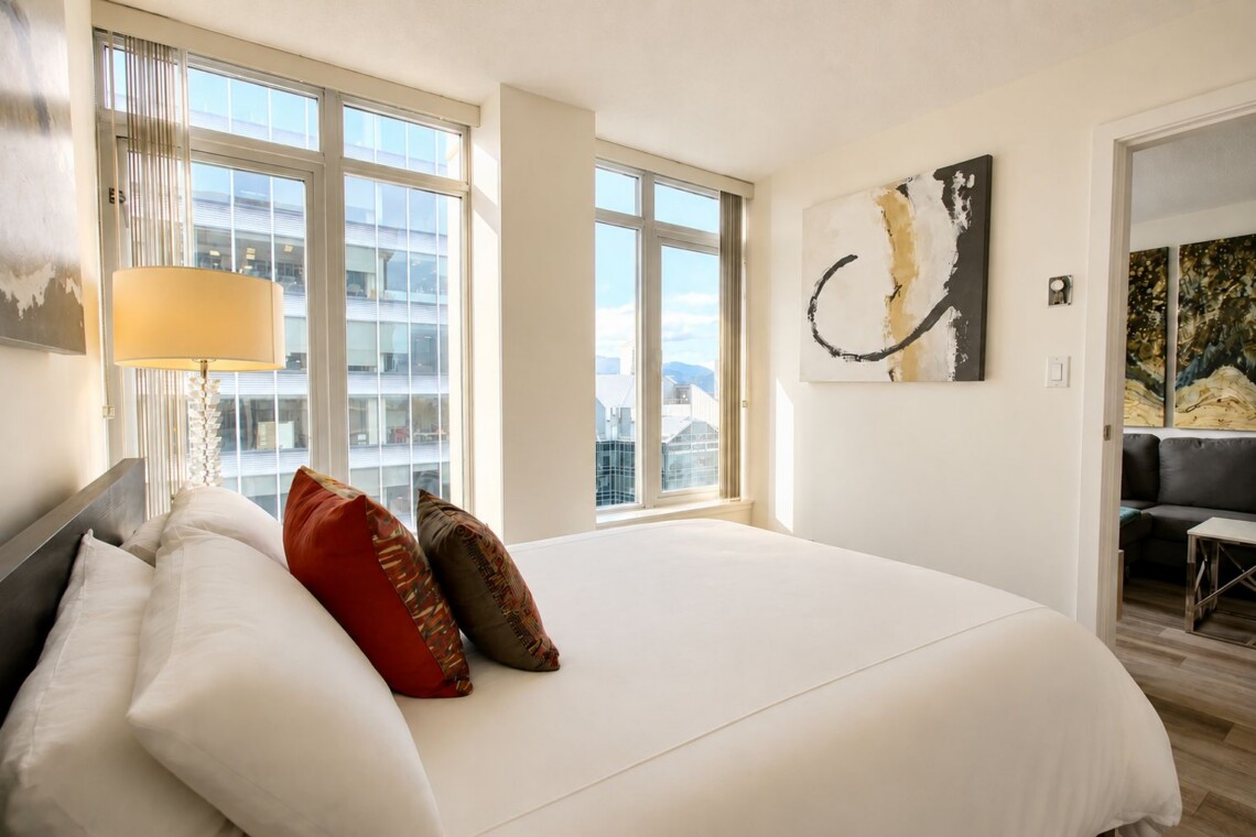 Bright hotel bedroom with a large white bed, red and brown decorative pillows, and a glass-based lamp beside floor-to-ceiling windows overlooking the city.