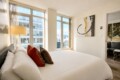 Bright hotel bedroom with a large white bed, red and brown decorative pillows, and a glass-based lamp beside floor-to-ceiling windows overlooking the city.