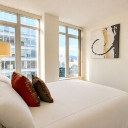 Bright hotel bedroom with a large white bed, red and brown decorative pillows, and a glass-based lamp beside floor-to-ceiling windows overlooking the city.
