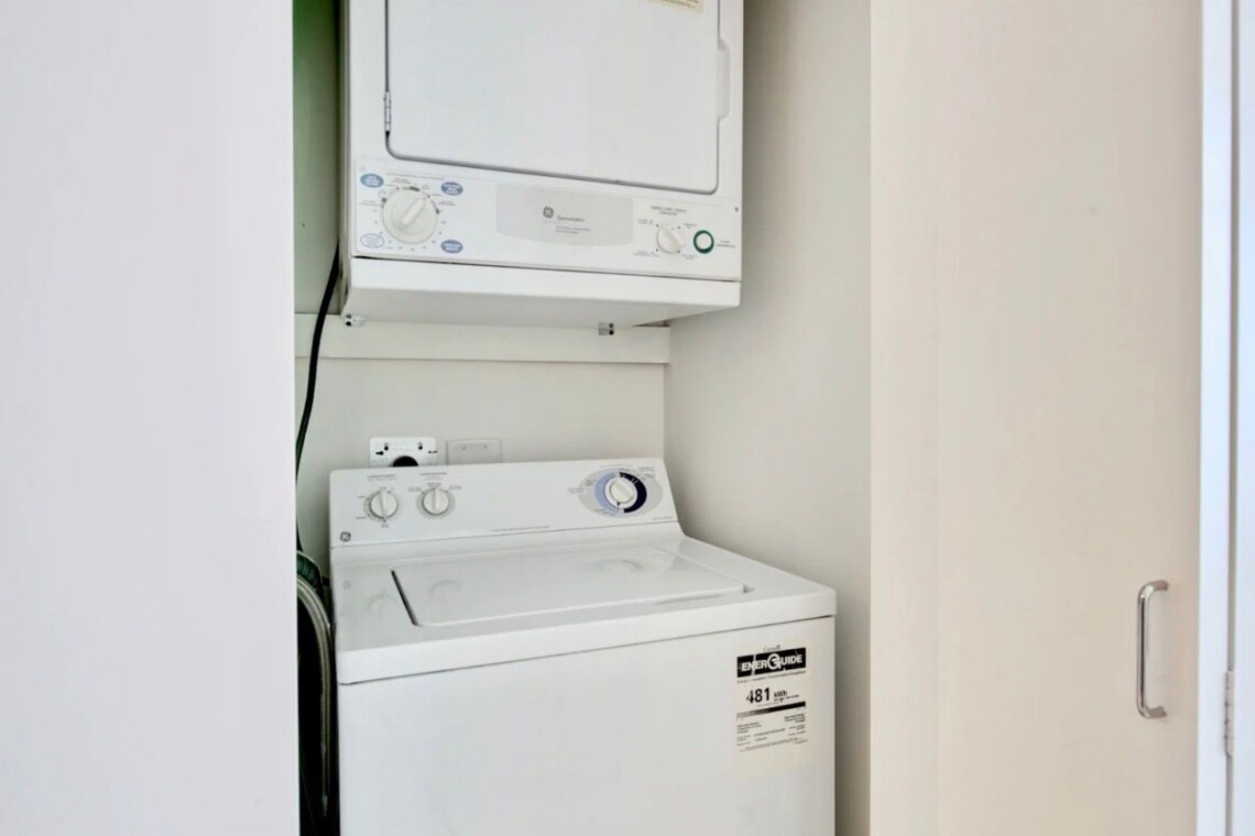 Stacked white washer and dryer in a narrow laundry alcove, with a doorway on the right side.