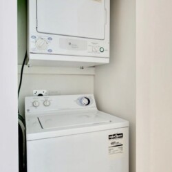 Stacked white washer and dryer in a narrow laundry alcove, with a doorway on the right side.