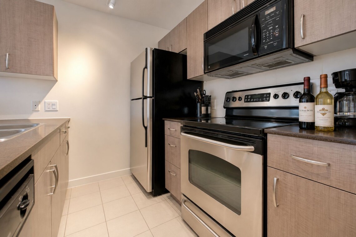 Compact kitchen with stainless steel fridge, four-burner stove, microwave, and wine bottles on the counter edge of a dark countertop area.