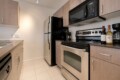 Compact kitchen with stainless steel fridge, four-burner stove, microwave, and wine bottles on the counter edge of a dark countertop area.