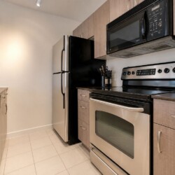 Compact kitchen with stainless steel fridge, four-burner stove, microwave, and wine bottles on the counter edge of a dark countertop area.