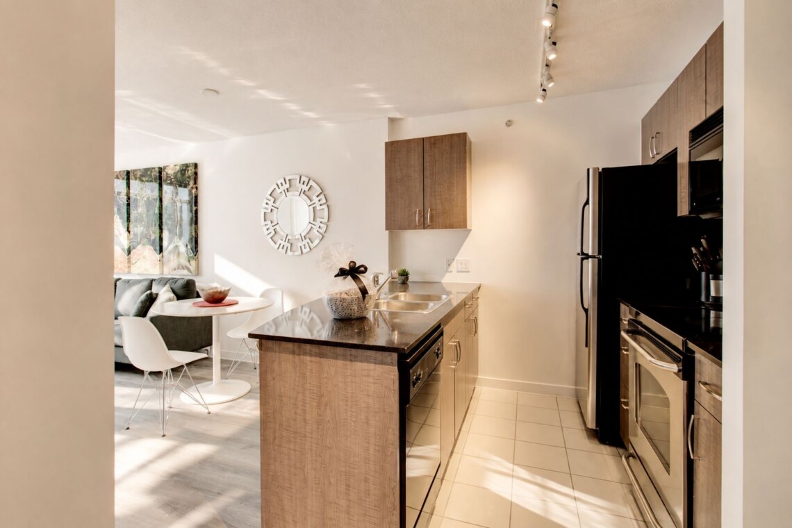 Open-concept kitchen with island, stainless steel appliances, and light wood cabinets; dining nook and living area visible in background.