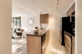 Open-concept kitchen with island, stainless steel appliances, and light wood cabinets; dining nook and living area visible in background.