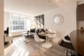 Open-plan living area with a gray sofa, white round dining table, wine bottles, and a large sunlit window