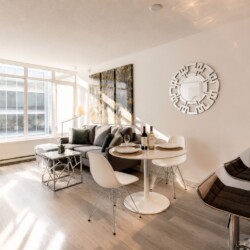 Open-plan living area with a gray sofa, white round dining table, wine bottles, and a large sunlit window
