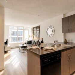 Open-concept kitchen with wood cabinets and island, set for two, adjacent living area with sofa and coffee table near a large window.