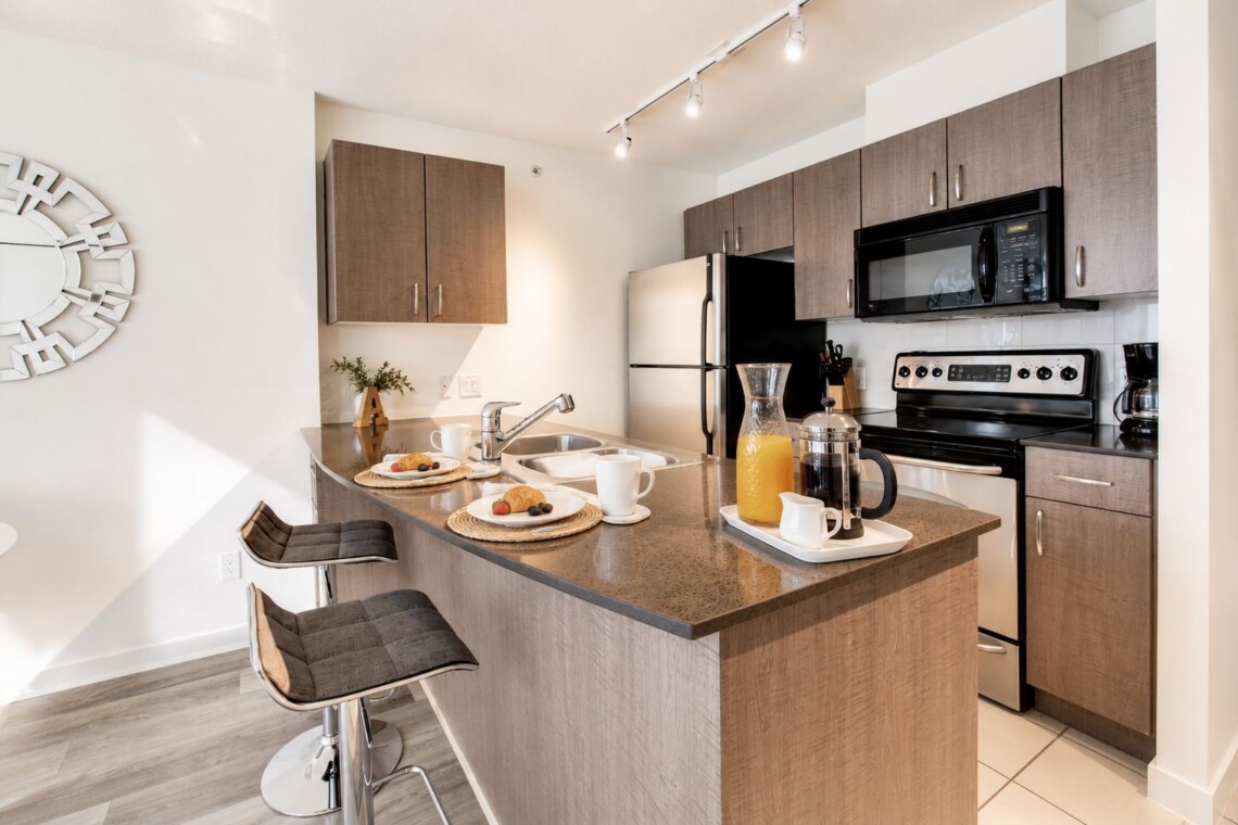 Modern kitchen with wood cabinets, an island, and a breakfast setup (pastries, coffee, and orange juice) for two under track lighting.