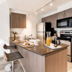Modern kitchen with wood cabinets, an island, and a breakfast setup (pastries, coffee, and orange juice) for two under track lighting.