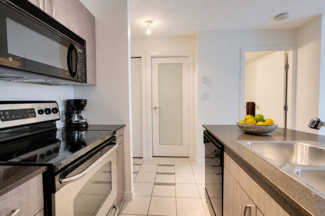Modern kitchen with stainless steel stove, microwave, and coffee maker along a gray countertop wall; island with double sink and a bowl of fruit ahead.