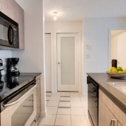 Modern kitchen with stainless steel stove, microwave, and coffee maker along a gray countertop wall; island with double sink and a bowl of fruit ahead.