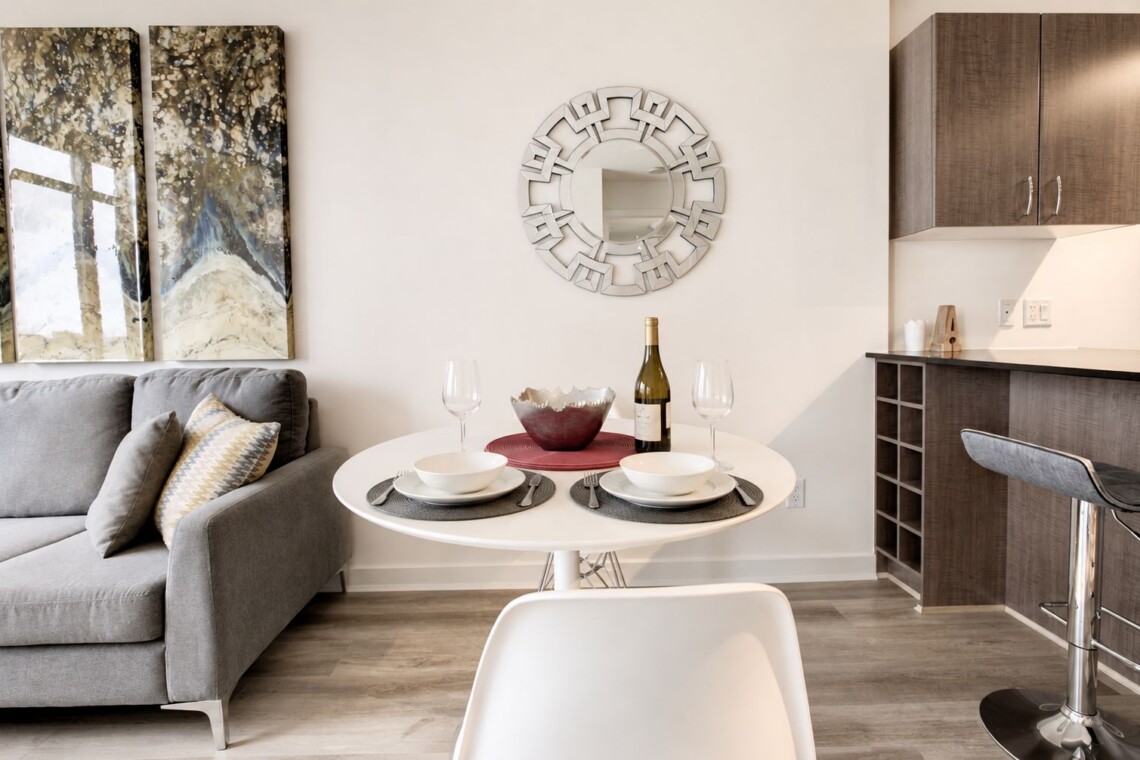 Modern living area with a round white dining table set for two, a wine bottle and glasses, and a gray sofa against a wall with abstract art.
