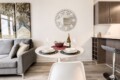 Modern living area with a round white dining table set for two, a wine bottle and glasses, and a gray sofa against a wall with abstract art.