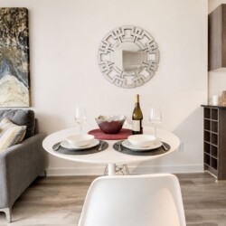 Modern living area with a round white dining table set for two, a wine bottle and glasses, and a gray sofa against a wall with abstract art.