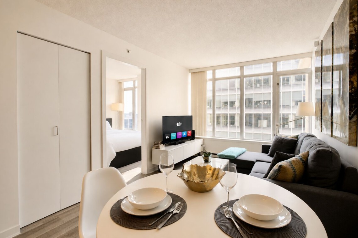 Bright open-plan living area with round dining table, gray sofa, large windows, and a TV on a white media console; bedroom doorway visible to the left.