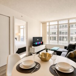 Bright open-plan living area with round dining table, gray sofa, large windows, and a TV on a white media console; bedroom doorway visible to the left.