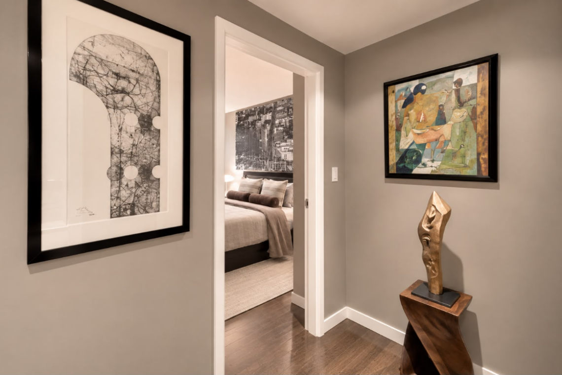 Hallway with gray walls, a large abstract black-and-white print on the left, a colorful painting on the right, and a sculptural wooden statue by a pedestal, leading to a bedroom.