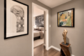 Hallway with gray walls, a large abstract black-and-white print on the left, a colorful painting on the right, and a sculptural wooden statue by a pedestal, leading to a bedroom.